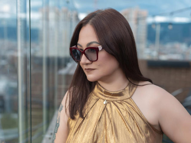 LadyNylonOh with brown hair, red-framed glasses, and a gold halter top, showing her tits and tattoo on her right arm, standing by a glass wall.