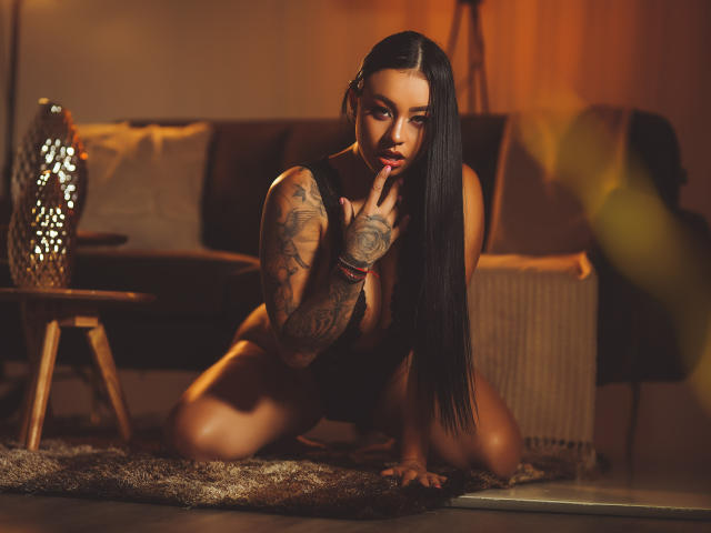 SarahMoonn with long black hair, tattooed arm, and dark lingerie, kneeling on a rug, finger in mouth, looking seductive, in a dimly lit room, showing her tits.