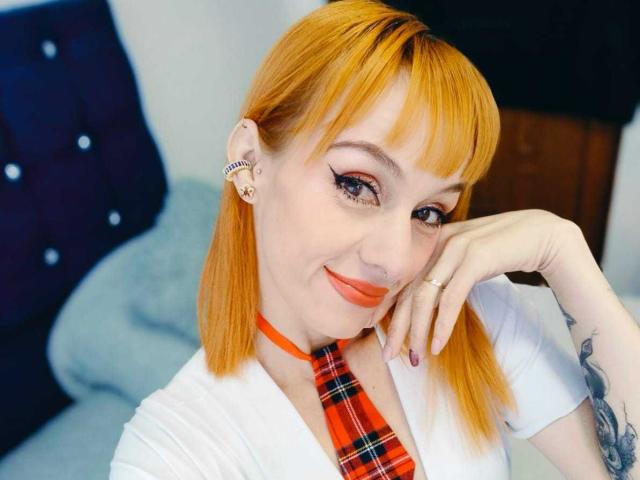 ScarlettN has orange hair, wearing a white shirt with a red plaid tie, smiling with red lipstick, right hand on cheek, visible tattoo on left arm, earrings, and ring.
