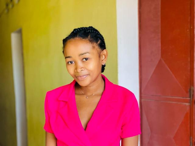 Samyra69 with medium brown skin, small breasts, black braided hair, wearing a bright pink V-neck shirt, smiling slightly, standing in front of a yellow and red wall.