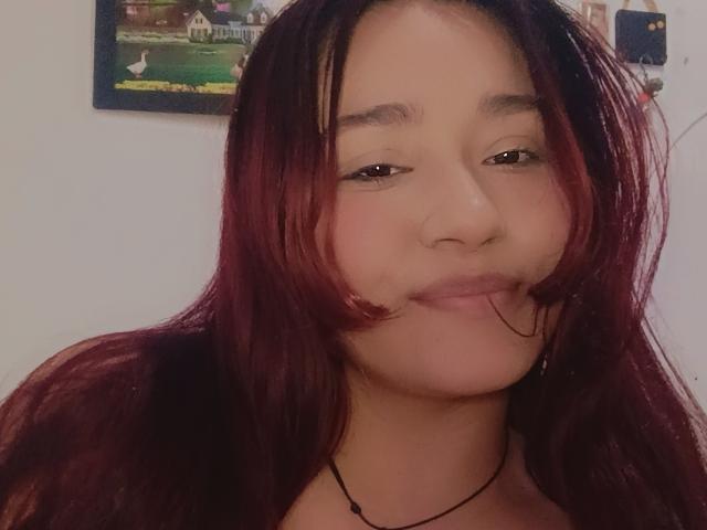 MaddieMoon ha i capelli rossi, gli occhi marroni e un leggero sorriso, con il labbro inferiore leggermente gonfio. Indossa una collana nera. Il suo viso è al centro della foto..