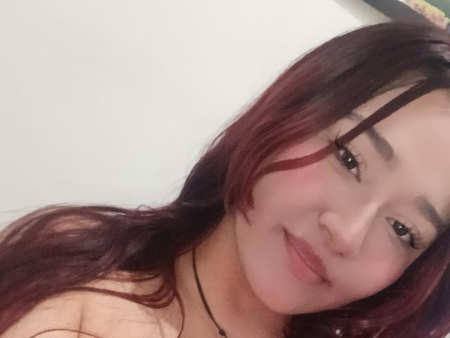 MaddieMoon mit rotem und schwarzem Haar, einem leichten Lächeln, einem schwarzen Choker und Wimpern, die ihr süßes Gesicht und ihre kleine Nase zeigen. Ihre Brüste sind leicht sichtbar..