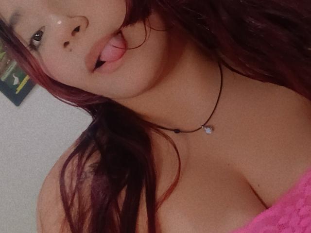 MaddieMoon con la lingua fuori, capelli rosso scuro, collana nera, top di pizzo rosa, che mostra il seno, pelle chiara e viso carino, in un selfie..