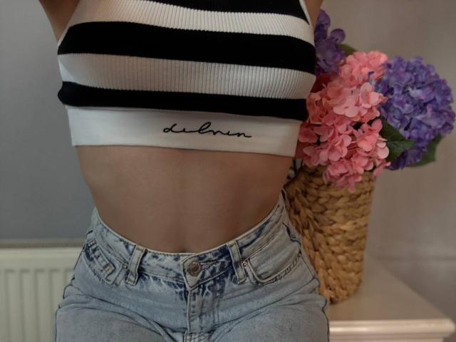 Amabel's striped crop top shows her small, perky tits, and she's wearing high-waisted, faded jeans with a visible waistband. A wicker vase with pink and purple flowers is nearby.