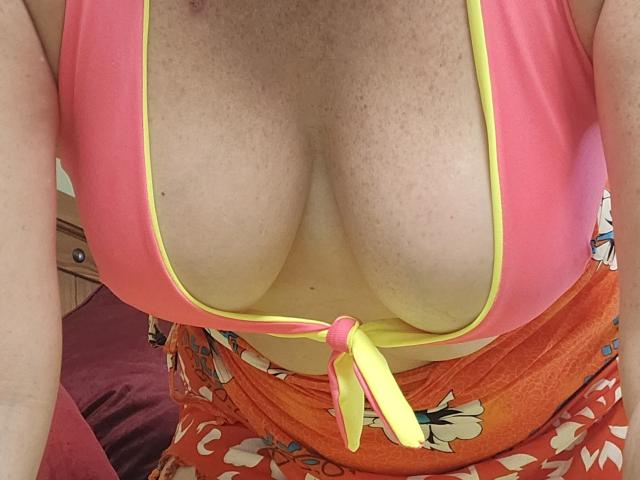 LylyCoquine69's big, freckled boobs in a pink and yellow top with a tied front, showing a lot of cleavage, wearing orange floral pants.