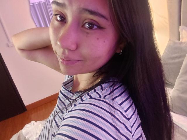 Karollteyes tem cabelos pretos longos, olhos castanhos, pequenas sardas e veste uma camisa listrada, com um sorriso discreto, a mão atrás da cabeça, em um quarto com iluminação suave..