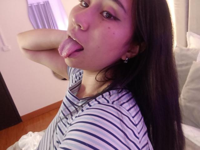 Karollteyes has long black hair, purple eyeliner, and is sticking out her tongue with her tongue covered in glitter. She's wearing a striped shirt, looking at the camera.