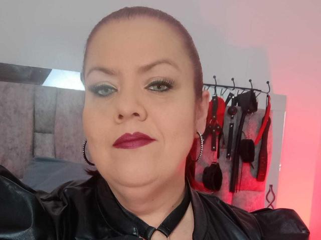 VanesaMoka with her hair pulled back, wearing black leather, red lipstick, and hoop earrings, has a confident expression, green eyeshadow, and a black choker in a sexy selfie.