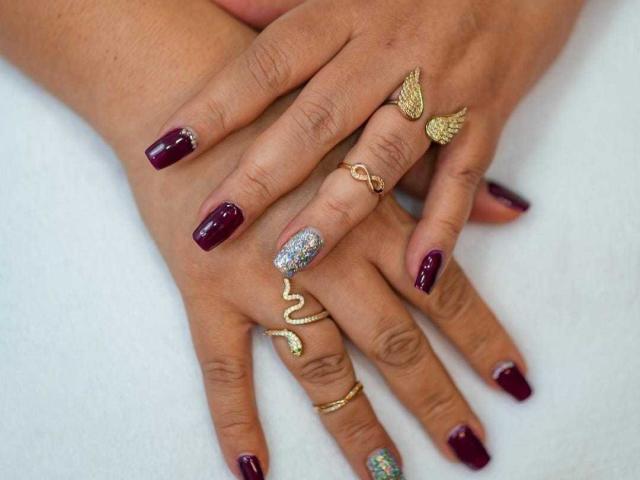VanesaMoka's hands with glossy dark red nails, one glittery silver nail, gold butterfly ring, snake ring, and gold wing ring. Her brown skin is smooth and elegant.