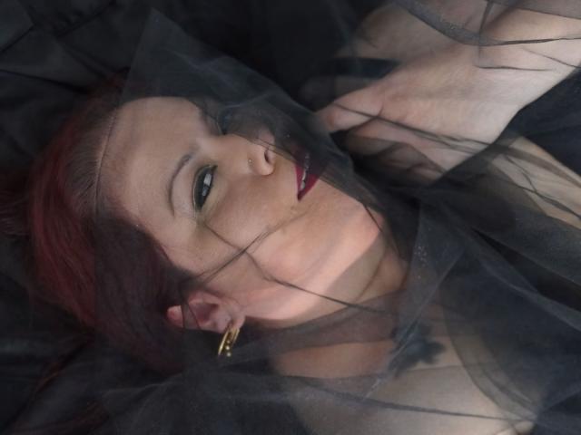 VanesaMoka with red hair, nose ring, and gold earrings, lying on black fabric, semi-transparent netting over her face, dark red lipstick, looking slightly to the side.