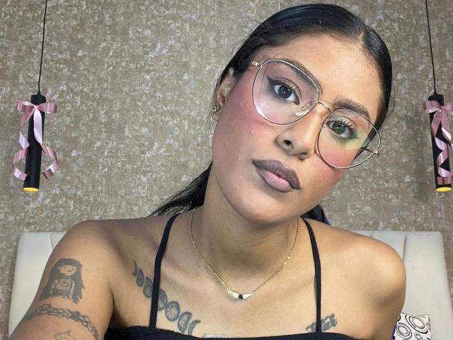 Babilett met een bril, een roze blush en donkere lipstick, die haar borsten met tatoeages laat zien, samen met een ketting en zwarte bandjes in een selfie. Ze heeft een serieuze uitdrukking..