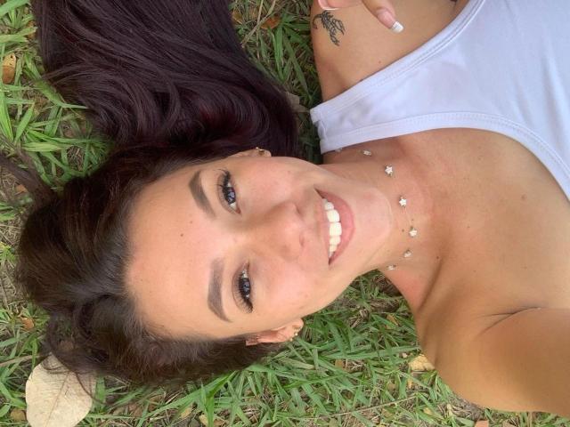 KinkyCharlottee lying on grass, smiling, brown eyes, white tank top, dark brown hair, small star necklace, tattoo on right shoulder, cute, pretty, sexy, visible collarbone, relaxed.