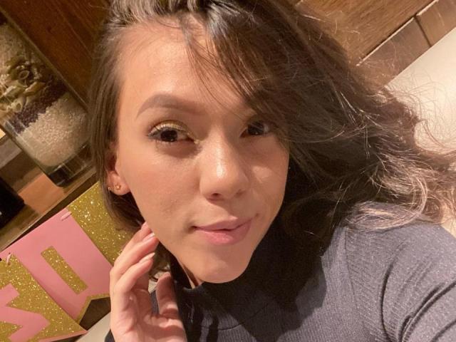 KinkyCharlottee has long, wavy brown hair, brown eyes, and a slight smile, wearing a gray turtleneck, with her right hand near her face in a selfie.