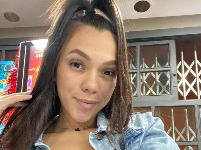 KinkyCharlottee has dark brown hair in a high ponytail, thick eyebrows, and a slight smile. She's wearing a denim jacket, and a hand is touching her hair.