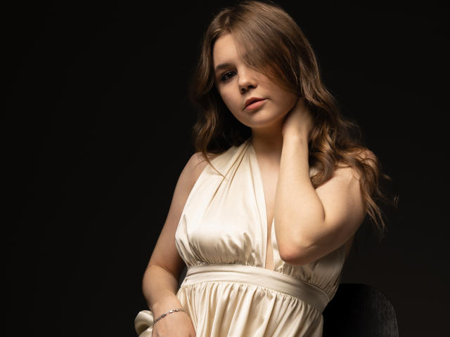 LeksaLove with wavy brown hair, wearing a silky, off-shoulder cream dress, touching her neck, dark background, subtle smile, soft lighting, small bracelet on left wrist.