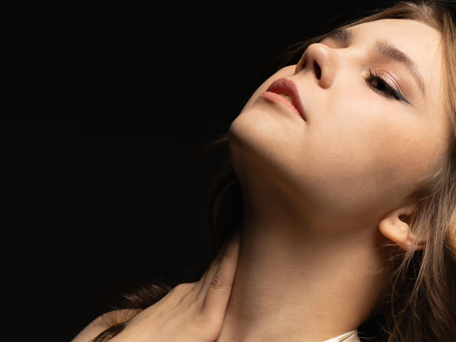 LeksaLove's face is tilted up, eyes half-open, with subtle makeup and brown hair. A hand gently touches her neck. Her lips are slightly parted. Dark background.