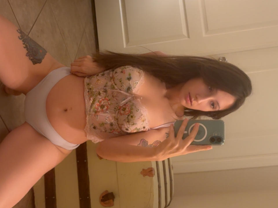 SkyllaCummies with long brown hair, wearing floral lace bralette and white panties, showing tattoos, holding phone, standing in bathroom, reflecting in mirror, showing her tits and ass.