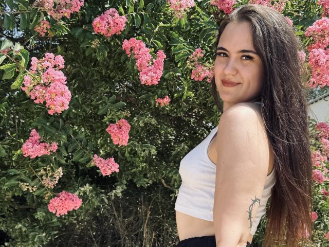 MarySweetSuprem with long brown hair, white tank top, black pants, small tits, tattoo on right arm, smiling at camera, surrounded by pink flowers, bright daylight.