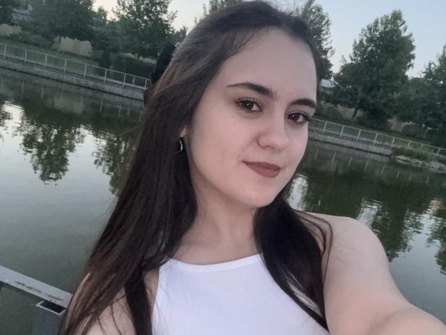 MarySweetSuprem has pale skin, long dark brown hair, and is wearing a white sleeveless top, showing her small tits in a selfie by a calm river.