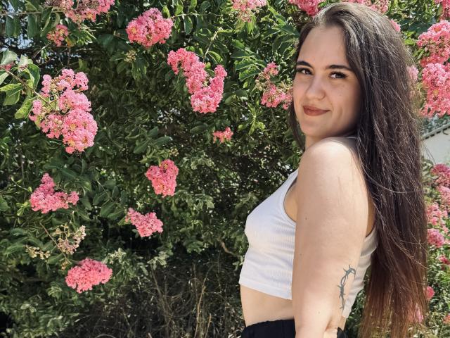 MarySweetSuprem with long brown hair, white crop top, black pants, smiling, pink flower bush, small rose tattoo on right arm, standing outdoors, sunlight, natural beauty.