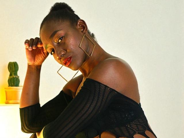 NahomyHawk with dark skin, red lipstick, and gold geometric earrings, wearing a black off-shoulder top, touching her head, and looking at the camera, with a cactus in the background.