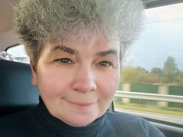 LiluYanata with a goofy, fluffy grey faux-hawk, smiling slightly, wearing a black turtleneck, sitting in a car with blurred greenery outside the window.