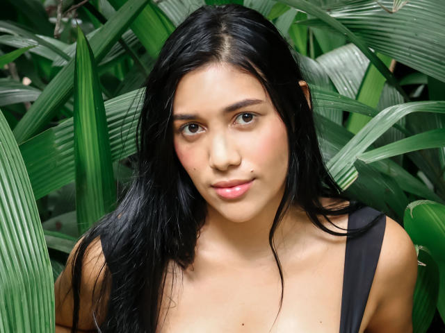 SaraRousse with long black hair, brown eyes, and pink lips, wearing a black top, showing her small tits, standing among green leaves.