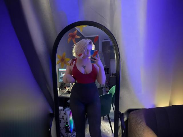 LimeCaty with blonde hair, red top, showing large boobs, black pants, holding phone in mirror, tattoo on chest, blue and purple lighting, standing in room, mirror reflection.