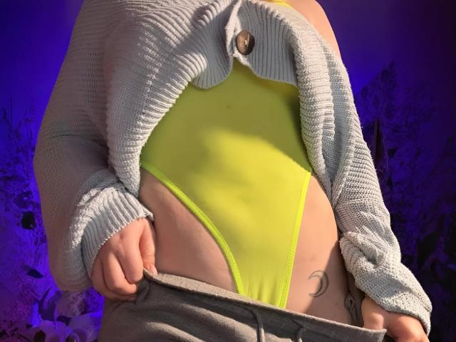 LimeCaty's yellow thong is visible under a gray knitted cardigan, with her hands pulling down gray pants, exposing a crescent moon tattoo on her right side.