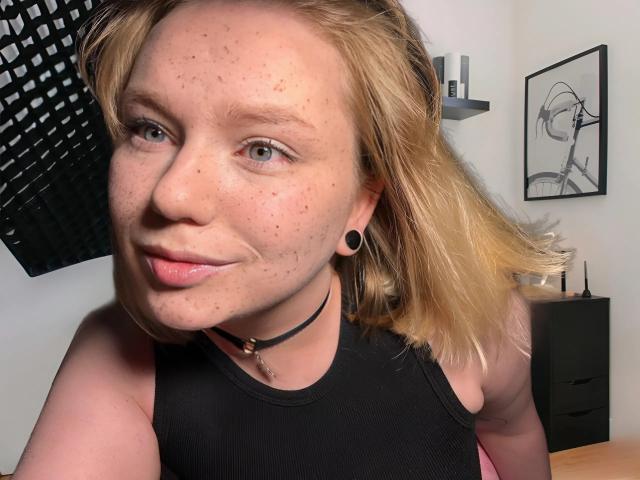 LimeCaty with blonde hair, black choker, black sleeveless top, black ear gauges, slight smile, freckles, and puffy lips, in a room with a black wall hanging.