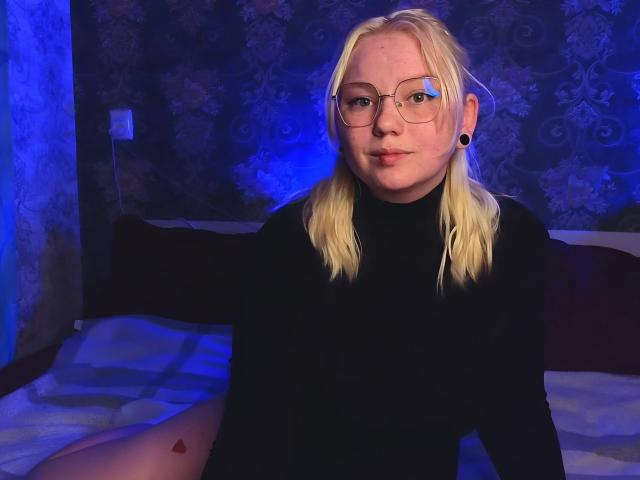 Blonde LimeCaty with pigtails, glasses, black turtleneck, and ear gauges, sitting on bed, blue lighting, small heart tattoo on left thigh, slightly chubby, pouty expression.