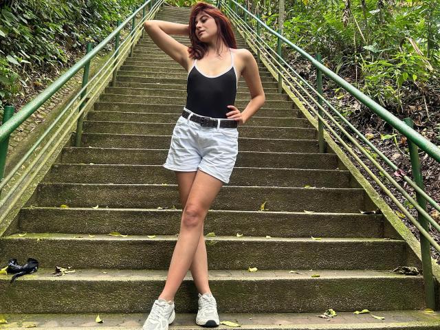SpencerLe stands on a mossy, green-railinged staircase, one hand behind her head. She's wearing a black tank top, white high-waist shorts, and white sneakers. Her red hair is loose, and she looks to the side with a relaxed expression. Her right leg is slightly bent, and her left leg is straight. The stairs have green railings and are surrounded by leafy greenery. A black shoe is on the bottom left. The composition is balanced and casual.