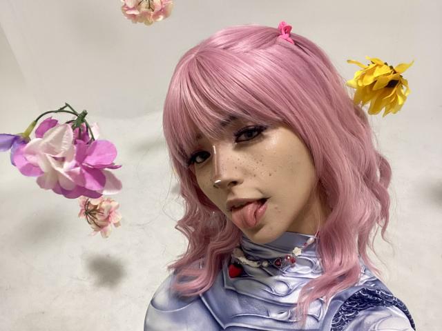CarlaMiceli with pink wig, sticking out tongue, wearing silver armor, and pink hair clips, has flowers floating around her head. She's cute with freckles and a playful expression.