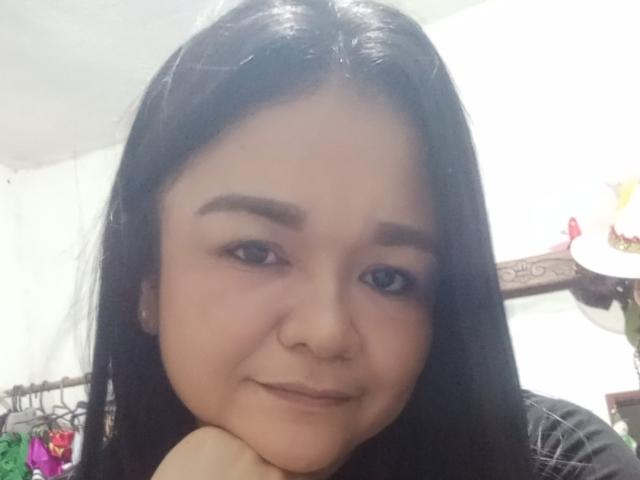TinaMarcia with long black hair, brown eyes, and a subtle smile, resting her chin on her hand, showing her nose and slightly raised eyebrows. Soft, natural makeup.