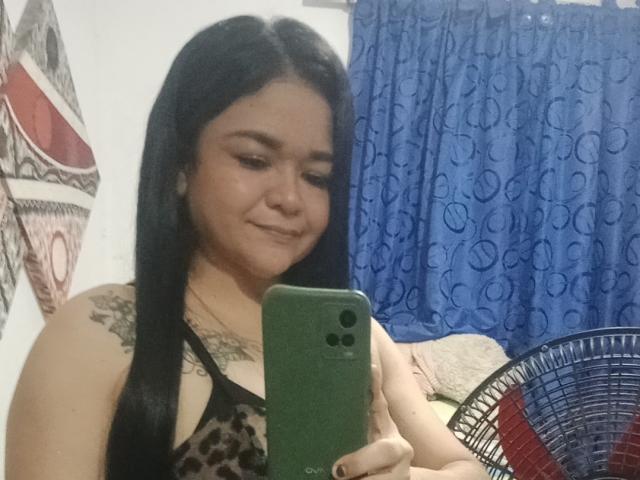 TinaMarcia with long black hair, tattoo on shoulder, smiling, wearing leopard print top, holding green phone, taking selfie in mirror, blue curtain background, fan on right.