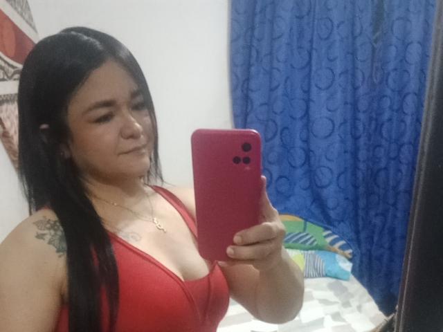 TinaMarcia with long black hair, red tank top, and small tattoo on right shoulder, holding red phone, taking selfie, showing her tits, and slight smirk. Bedroom setting.