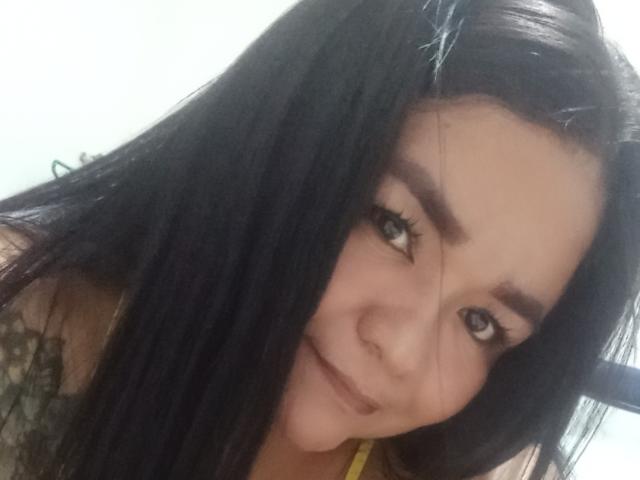 TinaMarcia has long black hair, thick eyebrows, and a slight smile. She's looking at the camera with her blue eyes. Her yellow top is visible.
