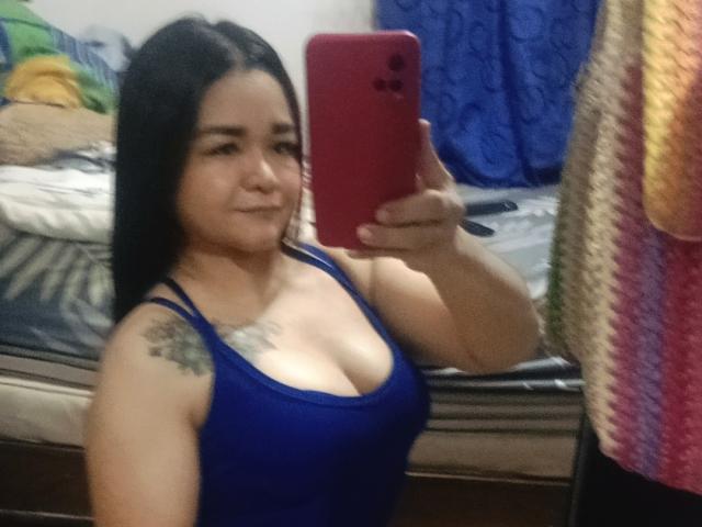 TinaMarcia with long black hair, blue tank top, and visible cleavage, holding a red phone, smiling, tattoo on left shoulder, standing in a bedroom. Boobs, phone, tattoo.