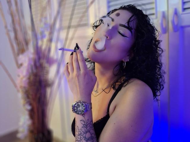 LilithFoxy with curly black hair, black lingerie, and tattooed arm, smoking a cigarette, eyes closed, nose pierced, blue light, and black nail polish, wearing a silver watch.