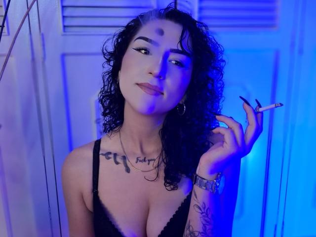 LilithFoxy with curly black hair, holding a cigarette, blue light, black lace bra, tattoos, cleavage, and a confident smile. Boobs, face, and a relaxed vibe.