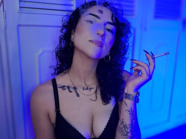 LilithFoxy with curly black hair, holding a cigarette, blue light on her face, wearing a black bra, tattoos on chest and arm, looking confident, medium tits.