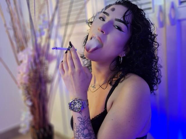 LilithFoxy with curly black hair, dark eyebrows, and black eyeliner, sticking out her tongue while smoking a cigarette, wearing a black lace bra and a tattooed wrist.