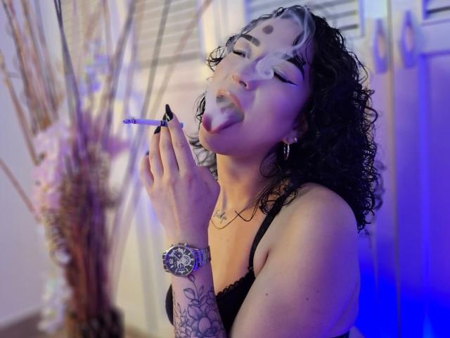 LilithFoxy with black curly hair, white and black makeup, tongue out, smoking, black lace bra, tattooed arm, silver watch, blue light, and black nail polish.