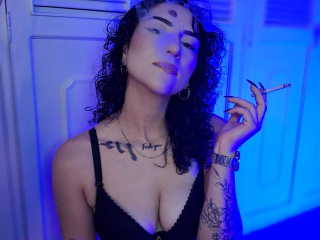 LilithFoxy with curly black hair, holding a cigarette, wearing black lace bra, showing tattoos on neck and arm, blue light, and hoop earrings, with a confident, slightly smug look.