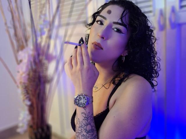 LilithFoxy with curly black hair, black lace bra, holding cigarette, blue light, watch on wrist, tattoo on arm, dark eyeliner, nose ring, and a third eye mark on forehead.