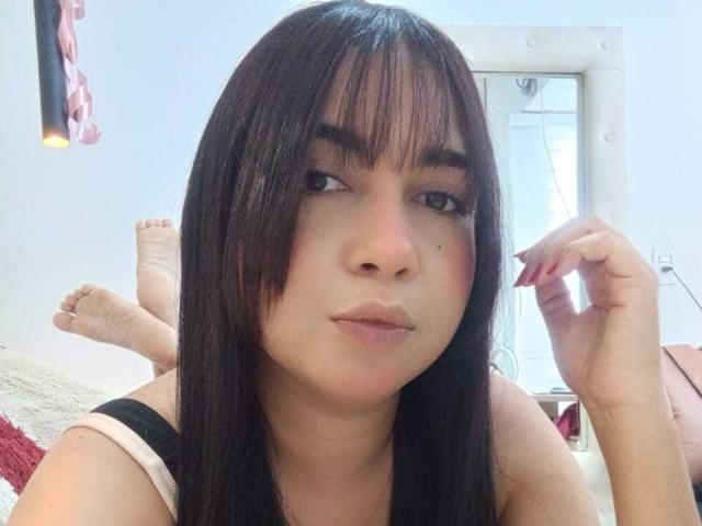 Alice Smity tiene el cabello negro y lacio con flequillo, ojos marrones y piel rosada. Está acostada en una cama, vestida con una blusa negra y rosa, con las piernas levantadas..