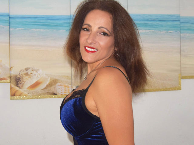 ReifeVivien-hot has medium-length brown hair and is smiling with red lipstick. She's wearing a blue velvet dress with black lace trim. Her skin is light, and she's looking over her shoulder. The background has a beach painting with seashells and blue-green ocean. Her earrings are small and silver. The image focuses on her upper body and face. Her hair is slightly messy. The painting adds a calm, coastal vibe.