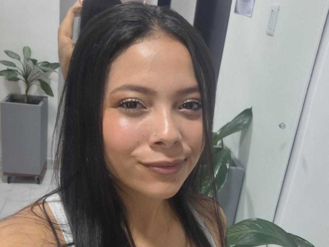 AnnieHill with shiny skin, black hair, brown eyes, and a subtle smile, wearing a white tank top, looking at the camera. Her ass is not visible.