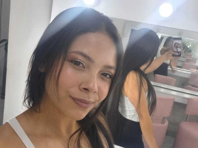 AnnieHill with dark hair, subtle makeup, nose piercing, white top, small smile, reflection in mirror showing back, taking selfie, pink chairs, bright lights, smooth skin.
