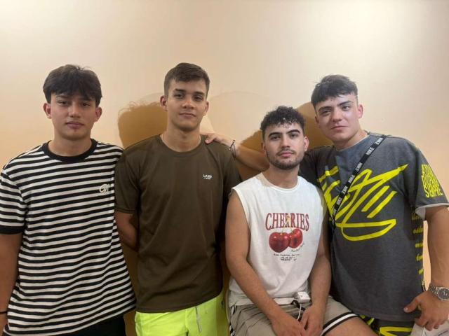 Four SpaceMen6s stand in a row, each wearing different shirts. The leftmost SpaceMen6 has a black and white striped shirt, the second from the left wears a brown shirt, the third SpaceMen6 has a white "Cherries" tank top, and the rightmost SpaceMen6 sports a gray shirt with yellow stripes. All have dark, curly hair. The third SpaceMen6 is sitting with his arm around the second SpaceMen6. Bright yellow shorts on the second SpaceMen6. Casual, indoor setting.